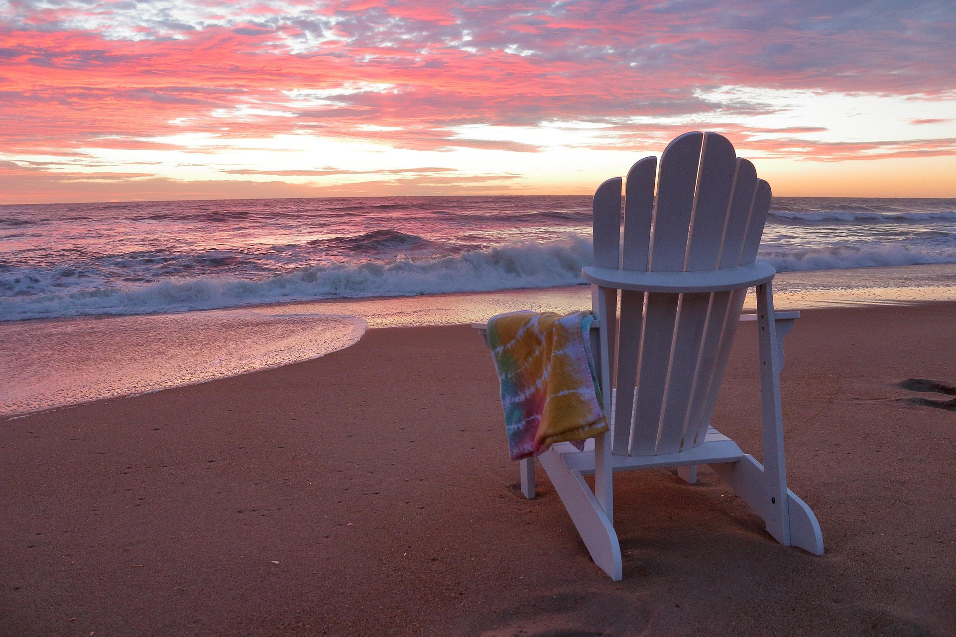 White Adirondack chair on a sandy beach overlooking the ocean at sunset.